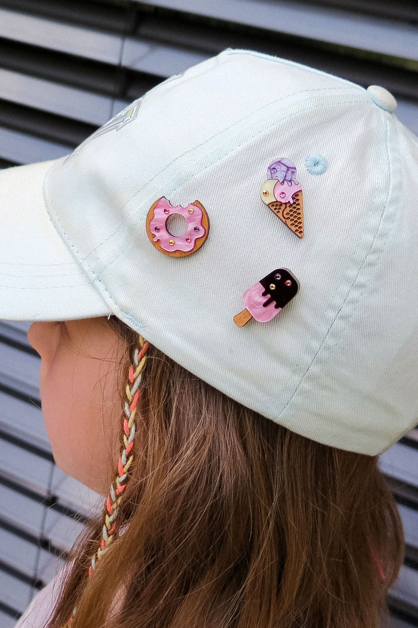 A child wearing a light-colored cap decorated with three cute handcrafted pins—a pink Donut Pin, an ice cream cone, and a popsicle—each adorned with colorful rhinestones. The background features horizontal gray metal slats.
