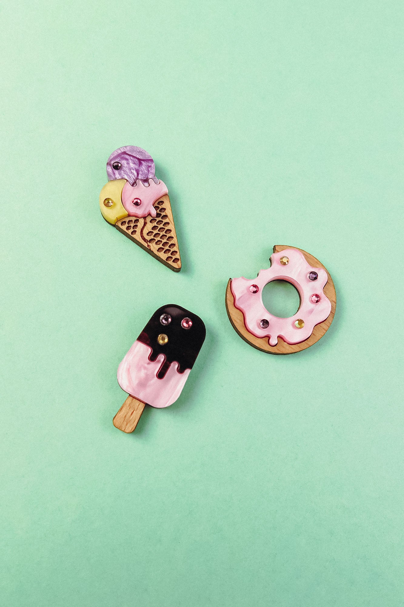 Three handcrafted clay pins on a mint green background: an ice cream cone, a Donut Pin with colorful rhinestones and sprinkles, and a pink-and-black popsicle.