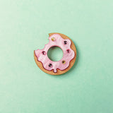 A handcrafted Donut Pin shaped like a donut with pink icing and colorful sprinkles, featuring a bite taken out of it, sits on a pastel mint green background.