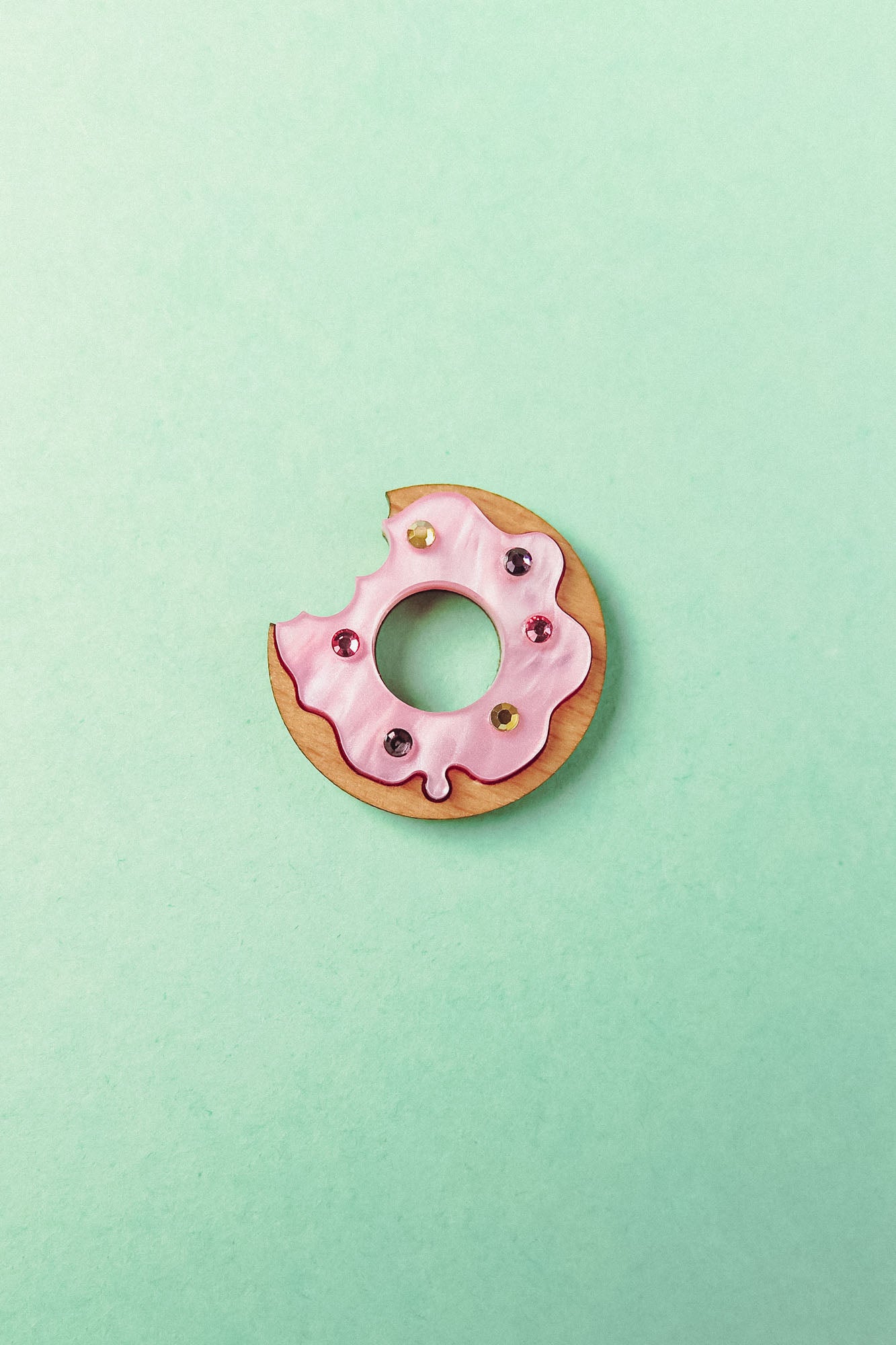 A handcrafted Donut Pin shaped like a donut with pink icing and colorful sprinkles, featuring a bite taken out of it, sits on a pastel mint green background.