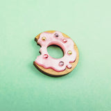 A handcrafted wooden Donut Pin with pink icing and colorful rhinestones, featuring a bite mark, sits on a mint green background.