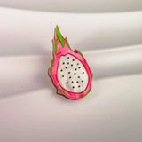 A colorful Dragon Fruit Pin, handcrafted with a white center and black seeds, is surrounded by a pink and green rind, bringing tropical charm to any outfit. Displayed against a wavy, white background.