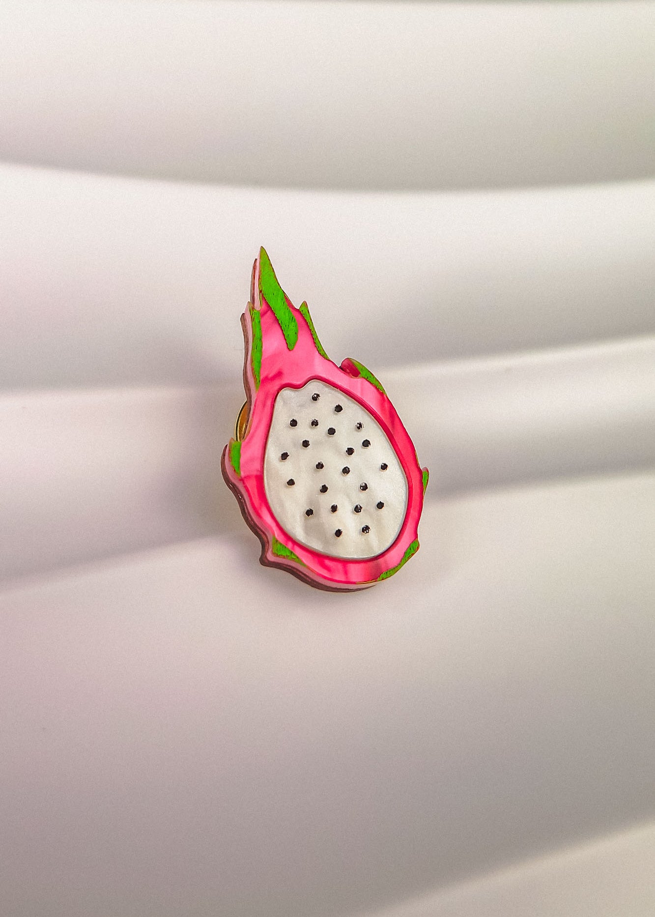 A colorful Dragon Fruit Pin, handcrafted with a white center and black seeds, is surrounded by a pink and green rind, bringing tropical charm to any outfit. Displayed against a wavy, white background.