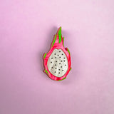 A handcrafted acrylic lapel pin that looks like a halved dragon fruit, with white center, black seeds, and pink and green edges, brings tropical charm against a light purple background.