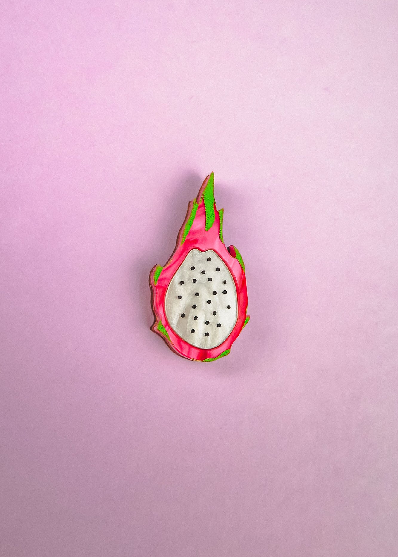 A handcrafted acrylic lapel pin that looks like a halved dragon fruit, with white center, black seeds, and pink and green edges, brings tropical charm against a light purple background.