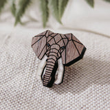 A handcrafted Elephant Pin with a geometric design and white tusks rests on textured beige fabric. Softly blurred green fern leaves appear in the background.