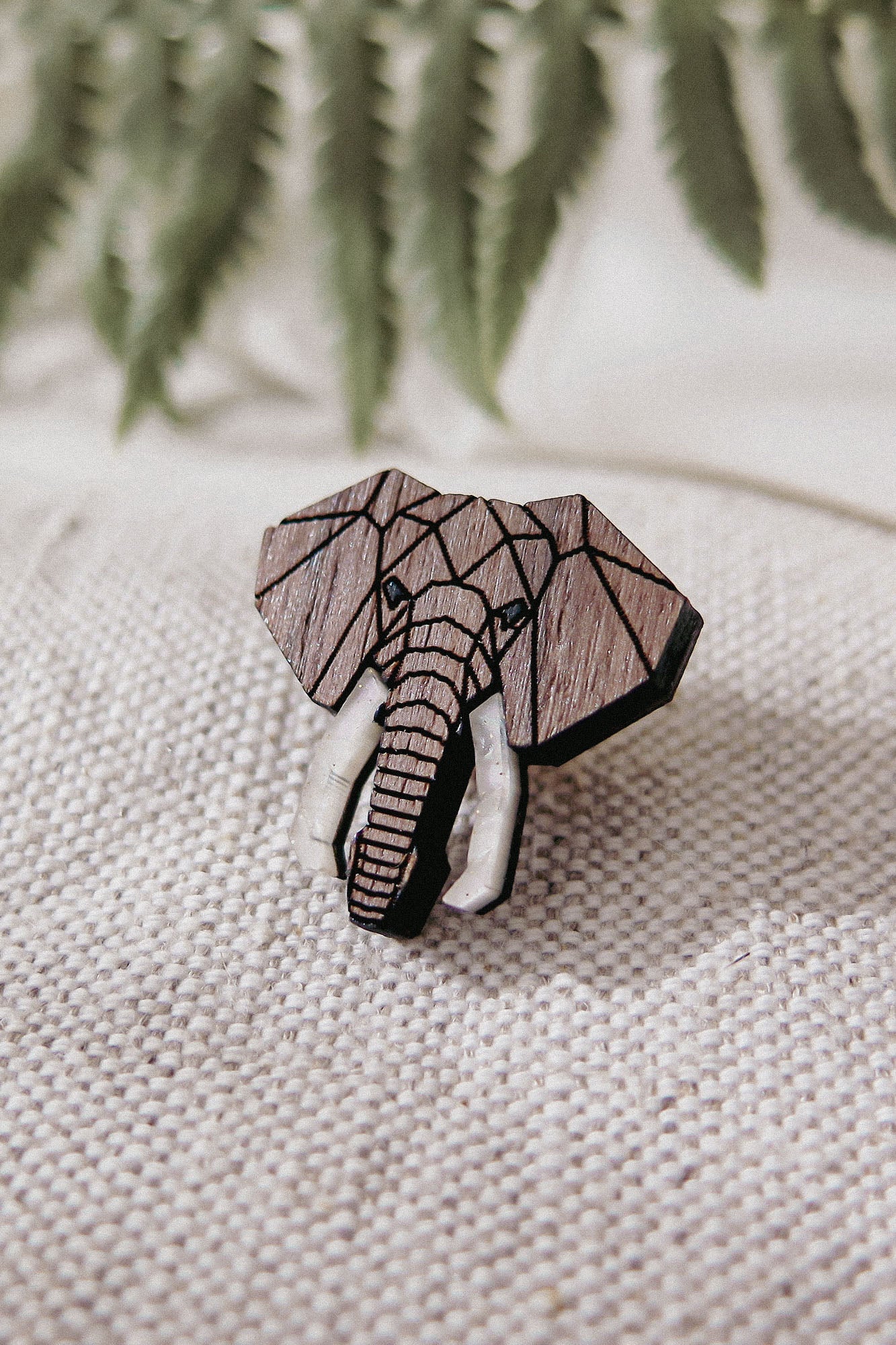 A handcrafted Elephant Pin with a geometric design and white tusks rests on textured beige fabric. Softly blurred green fern leaves appear in the background.