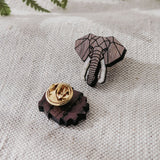 A handcrafted Elephant Pin shaped like an elephant’s head features a geometric design and white tusks, lying on textured fabric with its gold pin backing. Green fern leaves provide a vibrant background.