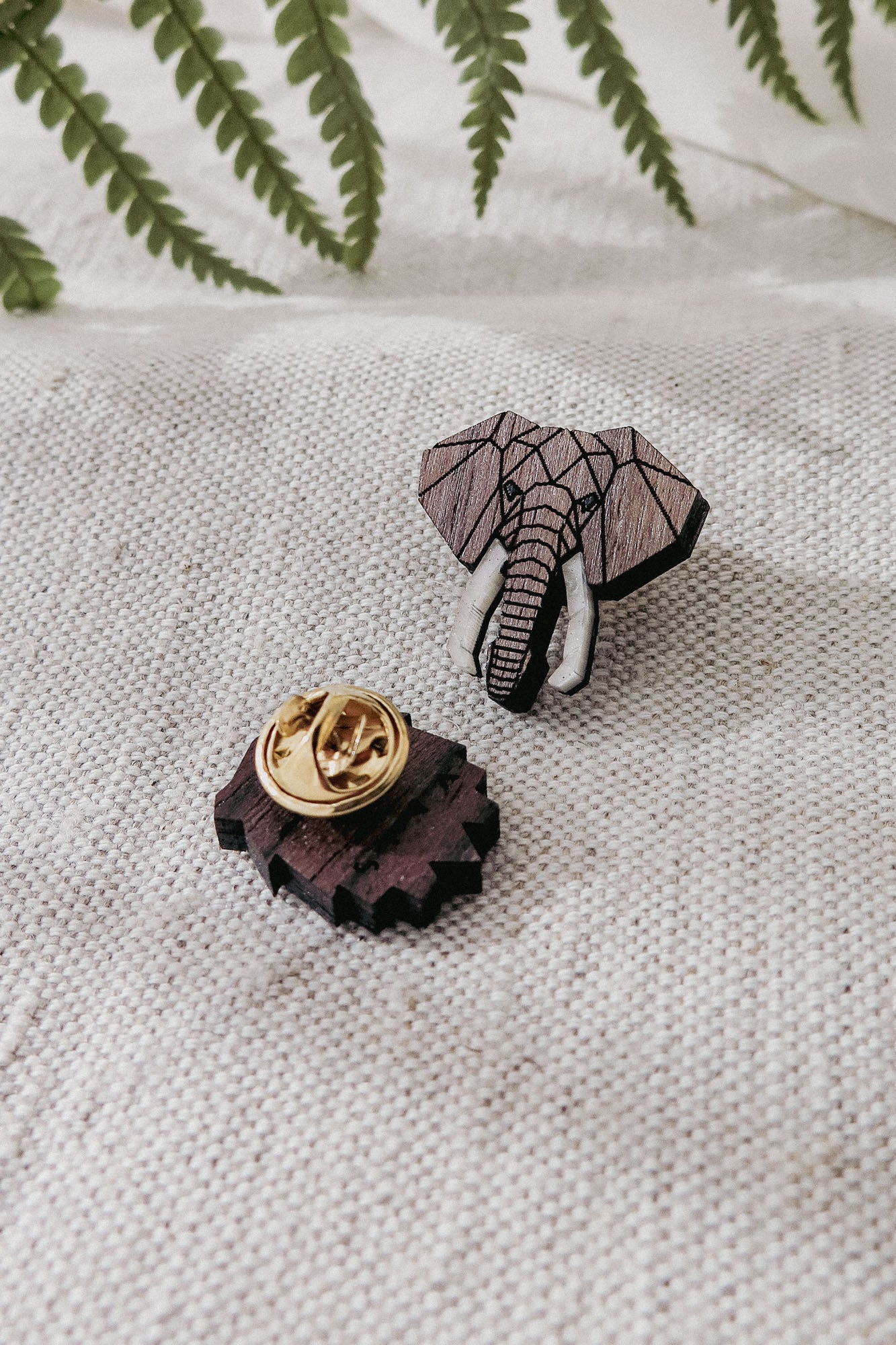 A handcrafted Elephant Pin shaped like an elephant’s head features a geometric design and white tusks, lying on textured fabric with its gold pin backing. Green fern leaves provide a vibrant background.