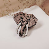 A handcrafted Elephant Pin shaped like an elephant’s head, featuring a striking geometric design, rests on a white surface with delicate dried flowers in the background.