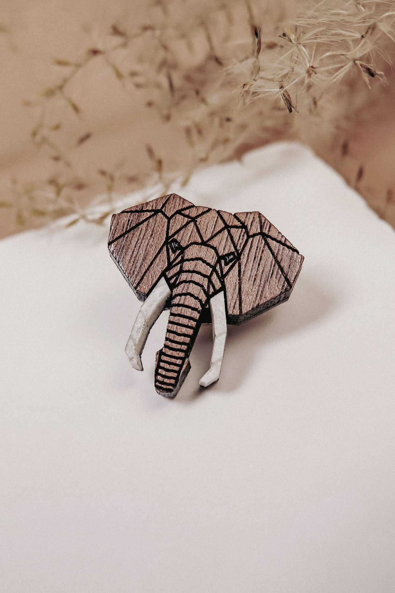A handcrafted Elephant Pin shaped like an elephant’s head, featuring a striking geometric design, rests on a white surface with delicate dried flowers in the background.