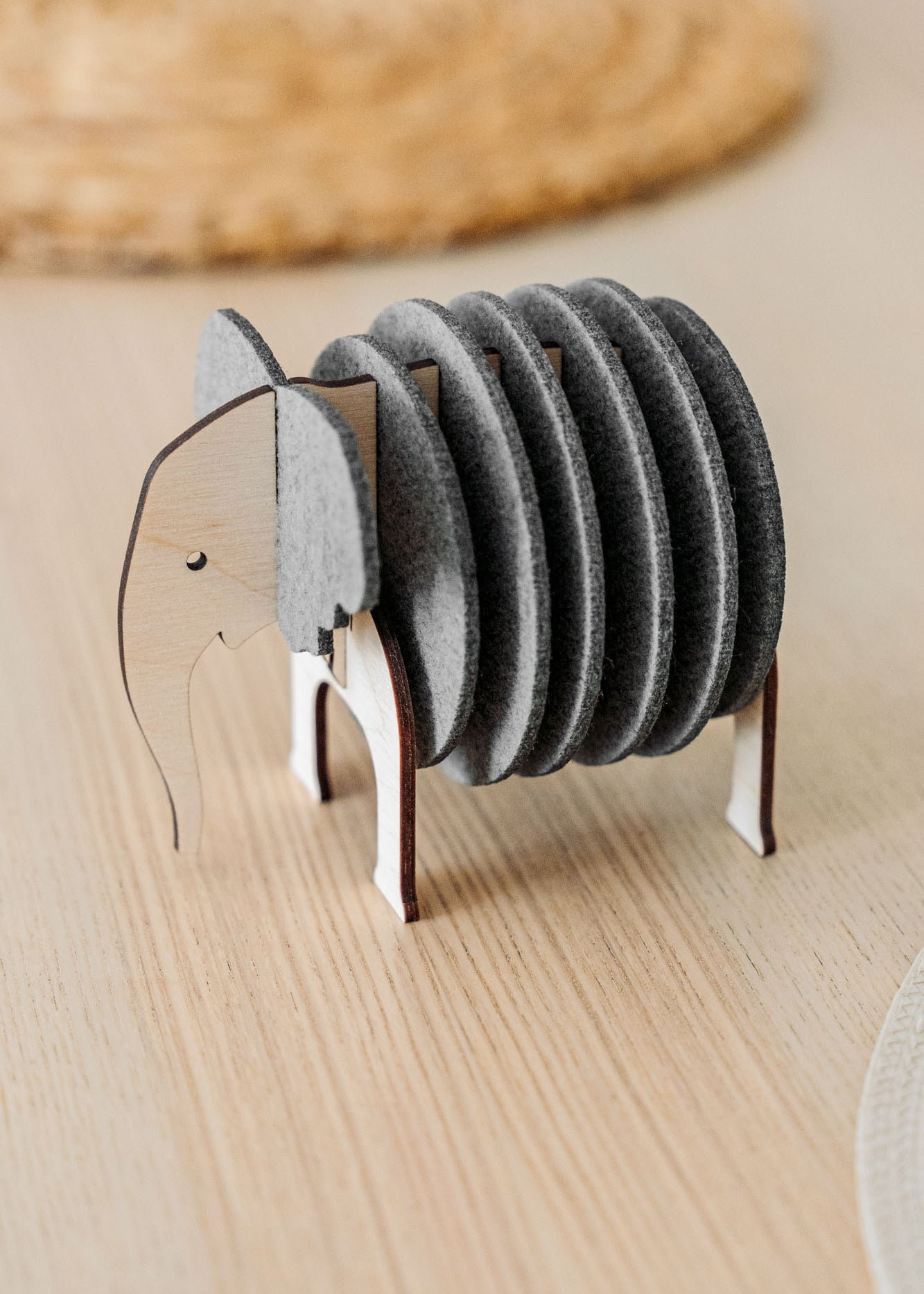 A set of round, gray Elephant Coasters is stacked vertically in a wooden holder shaped like an elephant, adding witty design and furniture protection to your light wood table.