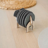 A set of round black Elephant Coasters is neatly arranged in a witty design sheep-shaped wooden holder, placed on a light wooden table with woven placemats in the background for stylish furniture protection.