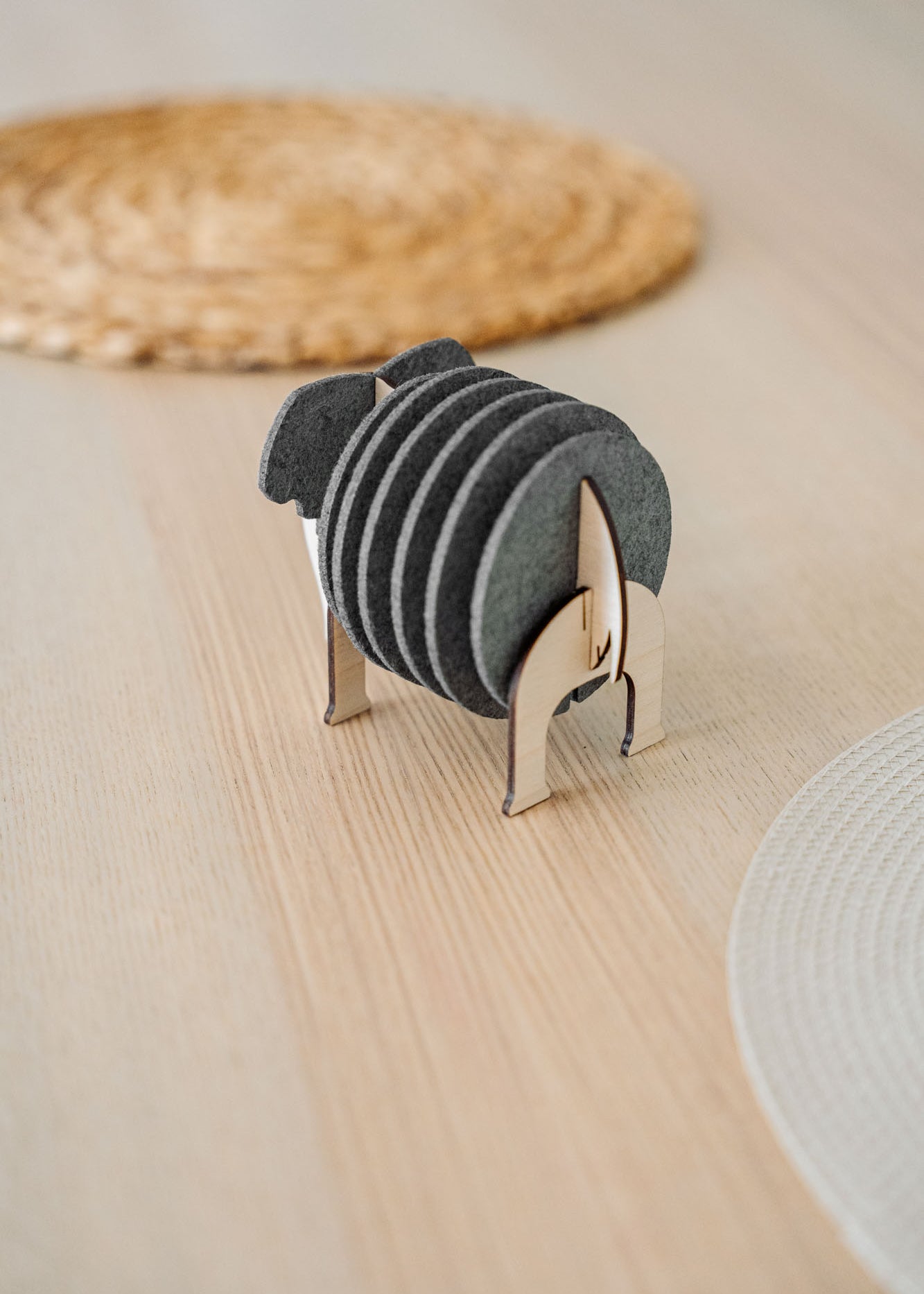 A set of round black Elephant Coasters is neatly arranged in a witty design sheep-shaped wooden holder, placed on a light wooden table with woven placemats in the background for stylish furniture protection.