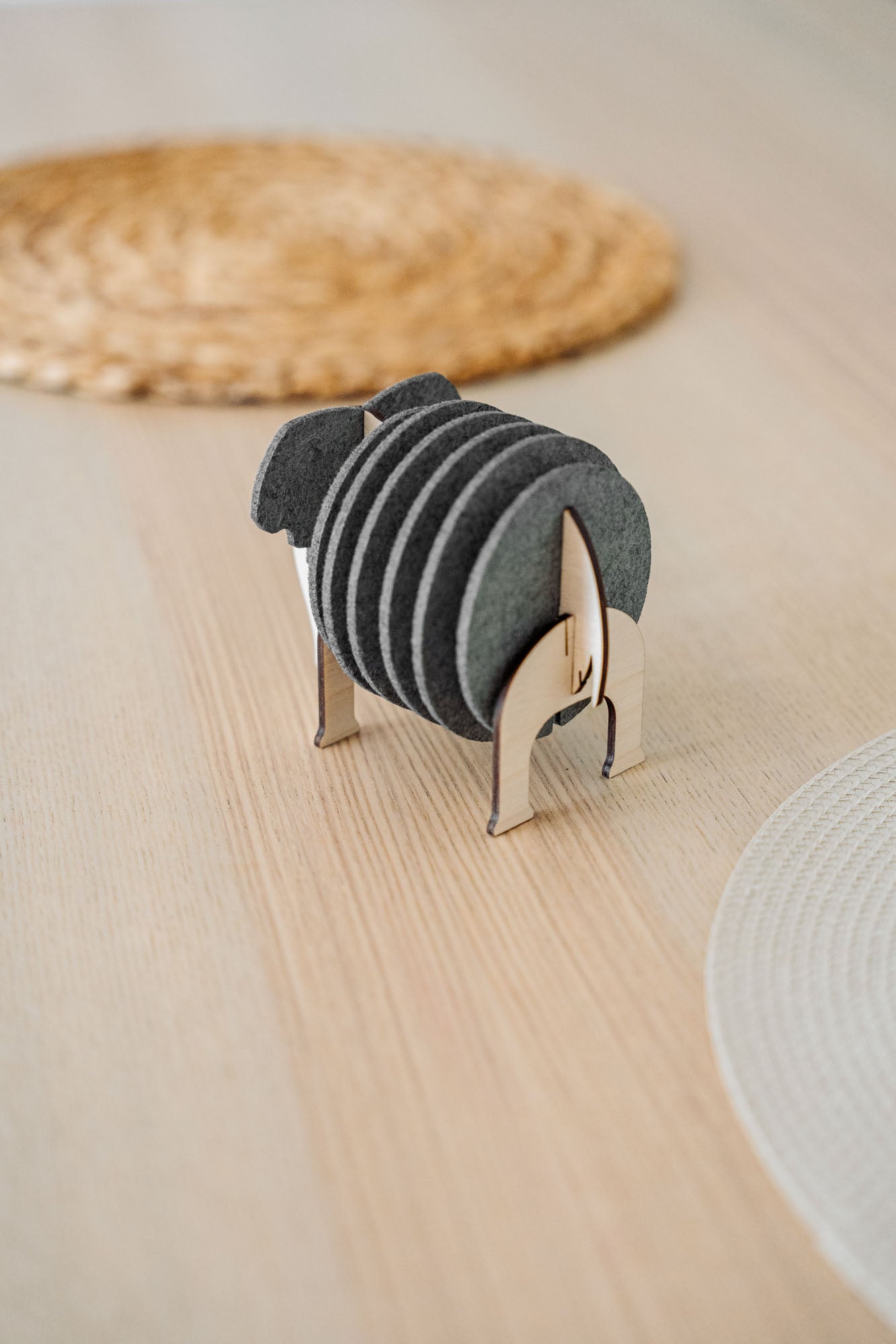 A set of round black Elephant Coasters is neatly arranged in a witty design sheep-shaped wooden holder, placed on a light wooden table with woven placemats in the background for stylish furniture protection.