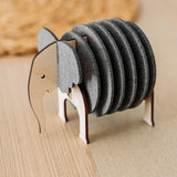 A wooden and felt Elephant Coasters holder with witty design stands on a wooden table, offering both charm and furniture protection, while a round woven placemat is blurred in the background.