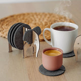 A set of Elephant Coasters with witty design offers stylish furniture protection, nestled in a wooden elephant-shaped holder on a wooden table beside a pink ceramic cup, white mug, and woven placemat.