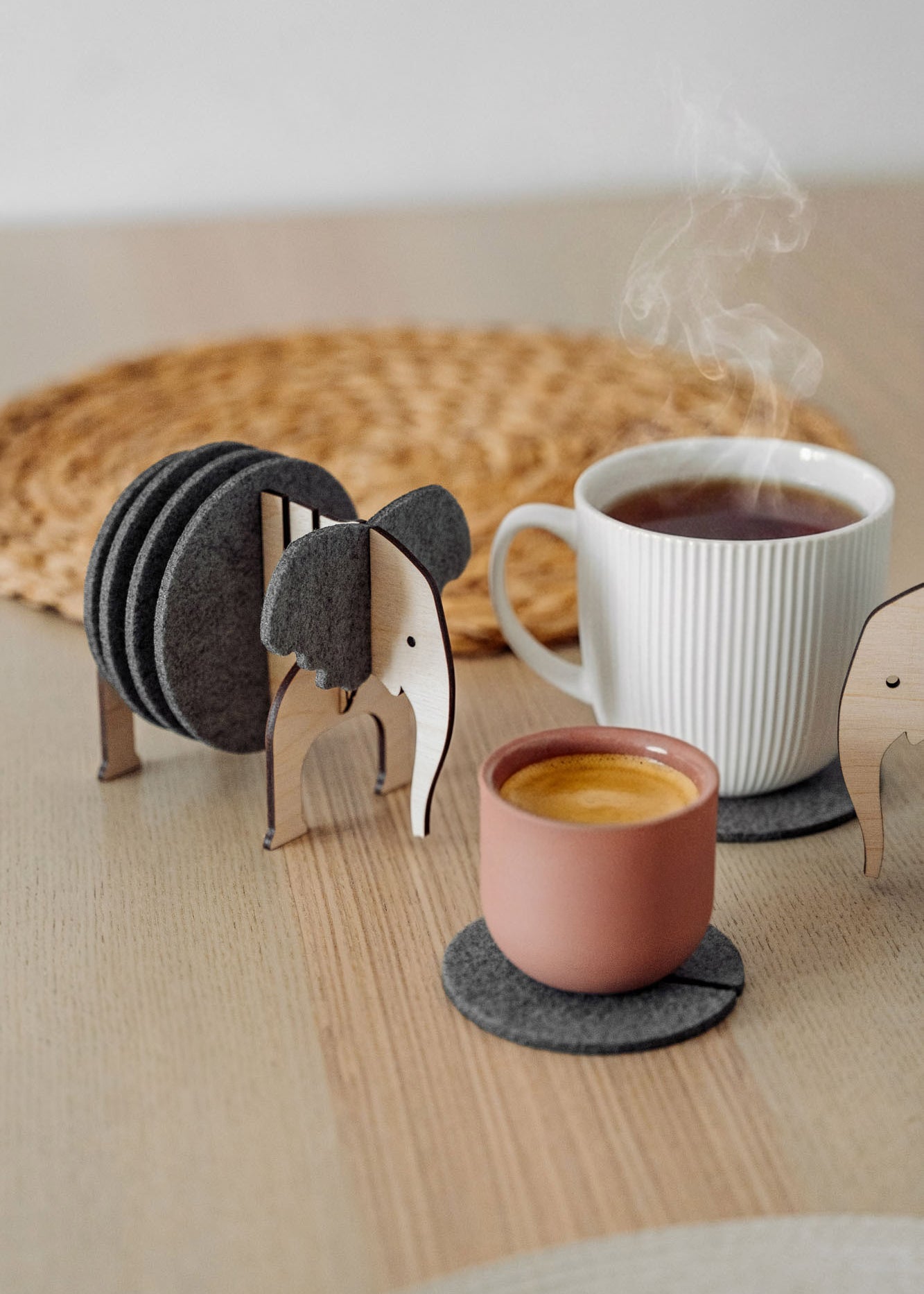 A set of Elephant Coasters with witty design offers stylish furniture protection, nestled in a wooden elephant-shaped holder on a wooden table beside a pink ceramic cup, white mug, and woven placemat.