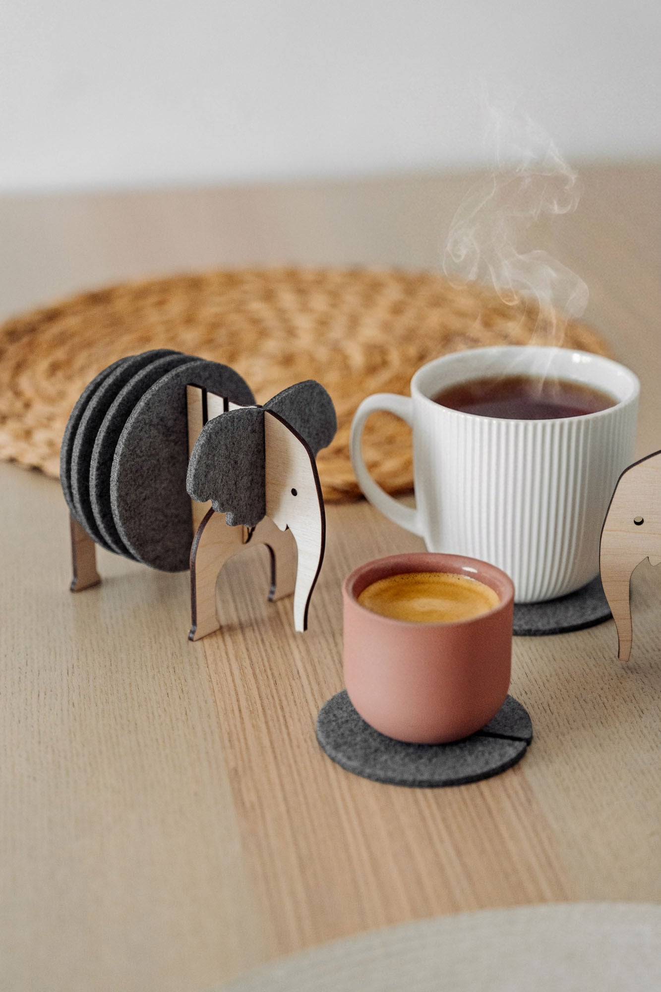 A set of Elephant Coasters with witty design offers stylish furniture protection, nestled in a wooden elephant-shaped holder on a wooden table beside a pink ceramic cup, white mug, and woven placemat.