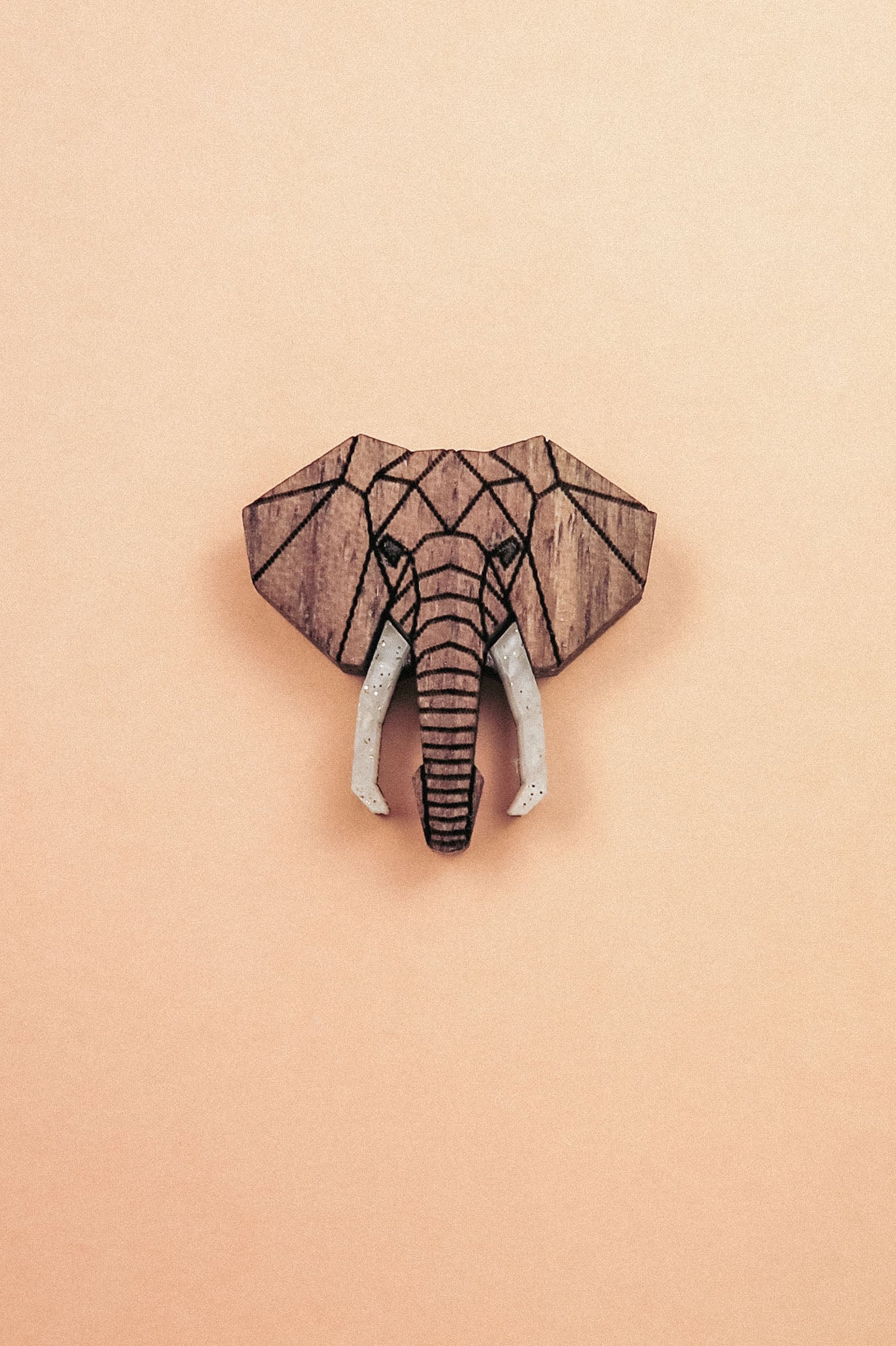 A handcrafted Elephant Pin showcases a geometric wooden carving of an elephants head with white tusks, centered on a light peach background.