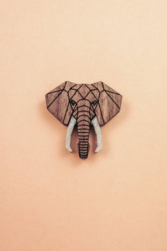 A handcrafted Elephant Pin showcases a geometric wooden carving of an elephants head with white tusks, centered on a light peach background.
