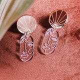 Silver handcrafted Face Earrings with abstract designs and round, reflective tops rest on a soft pink surface. A green and white leaf partially frames the image, adding artful sophistication to elevate your style.