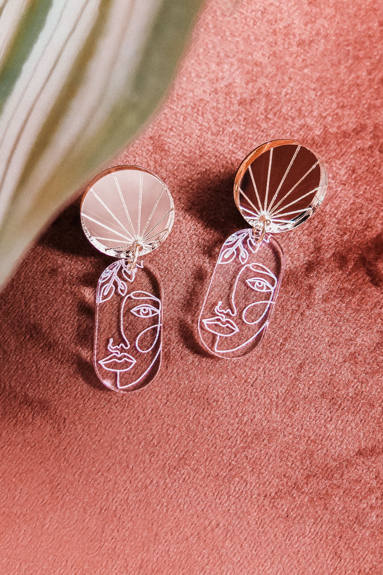 Silver handcrafted Face Earrings with abstract designs and round, reflective tops rest on a soft pink surface. A green and white leaf partially frames the image, adding artful sophistication to elevate your style.