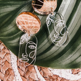 Gold and clear handcrafted Face Earrings with abstract faces rest on a large green leaf atop a woven straw mat, adding artful sophistication to elevate your style.