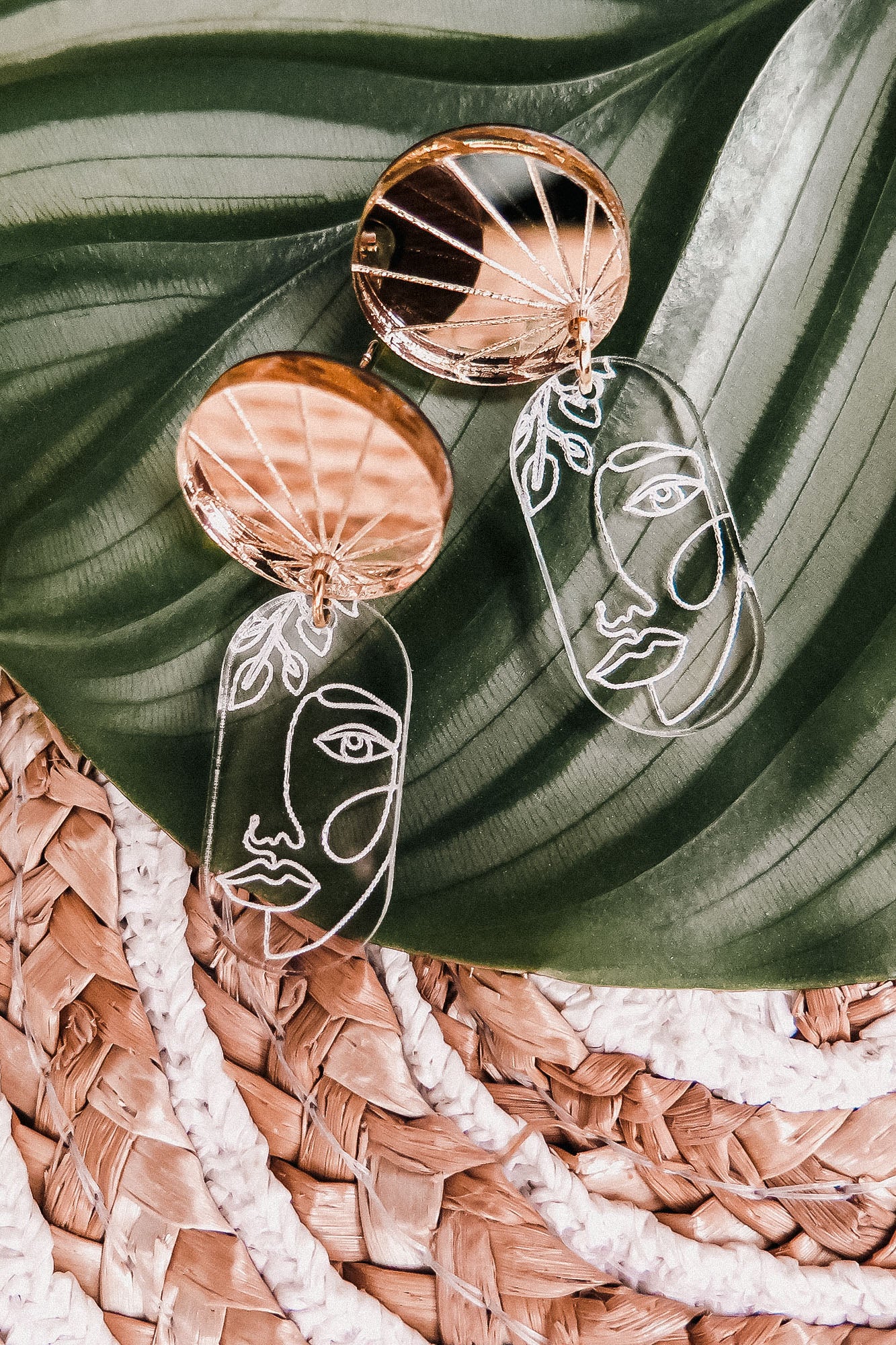 Gold and clear handcrafted Face Earrings with abstract faces rest on a large green leaf atop a woven straw mat, adding artful sophistication to elevate your style.