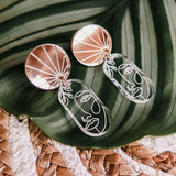 A pair of handcrafted Face Earrings with gold circular tops and abstract wire faces hang below, displayed on a green tropical leaf with a woven surface underneath to elevate your style with artful sophistication.