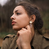 A woman with dark hair tied back gazes thoughtfully to the side outdoors, wearing a green jacket and handcrafted Face Earrings with a dangling design—an artful sophistication that gently elevates your style. Her hands are clasped near her chin.