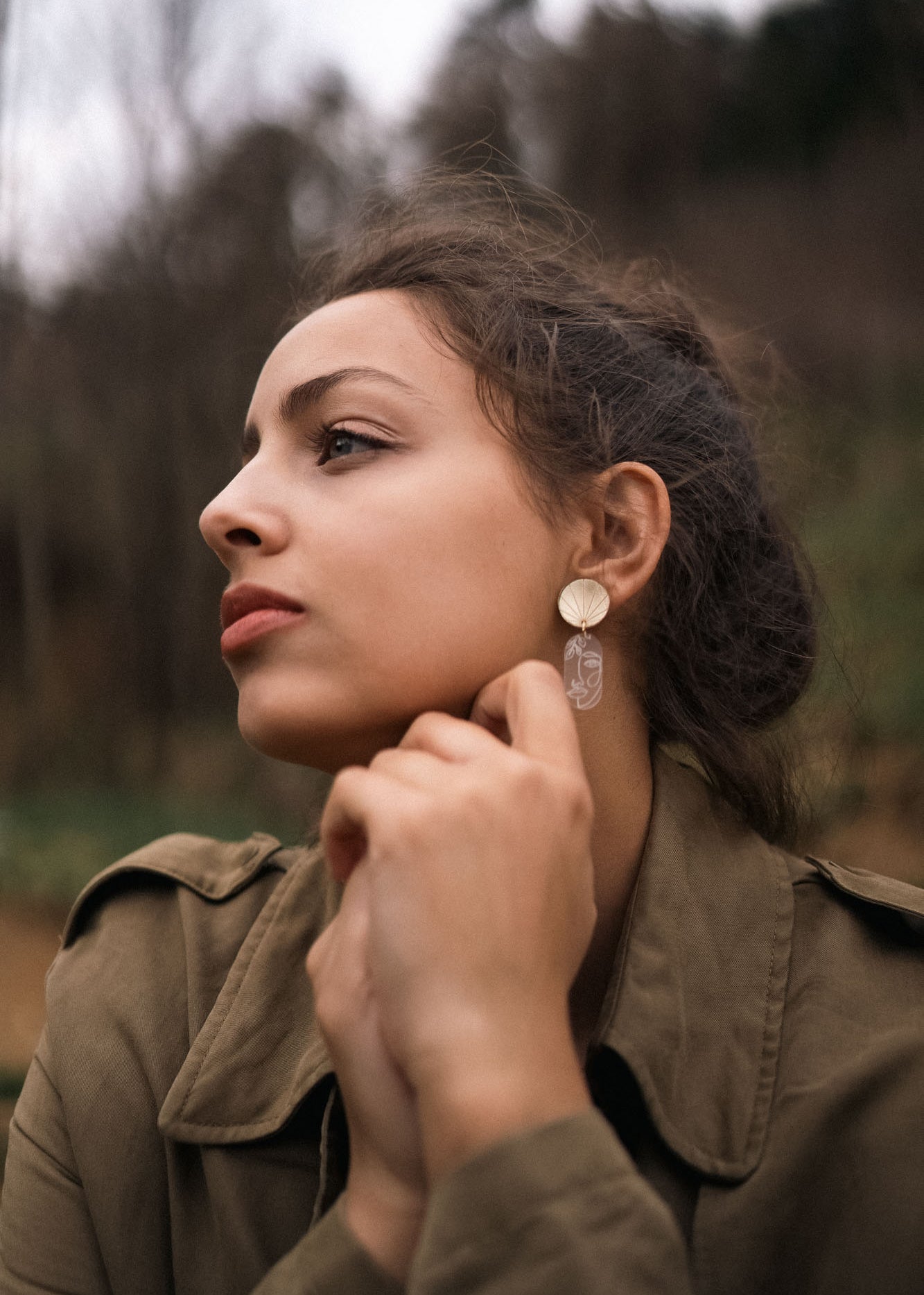 A woman with dark hair tied back gazes thoughtfully to the side outdoors, wearing a green jacket and handcrafted Face Earrings with a dangling design—an artful sophistication that gently elevates your style. Her hands are clasped near her chin.