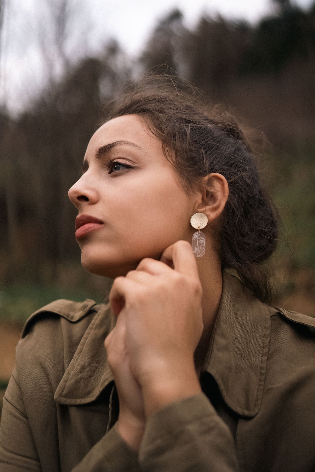 A woman with dark hair tied back gazes thoughtfully to the side outdoors, wearing a green jacket and handcrafted Face Earrings with a dangling design—an artful sophistication that gently elevates your style. Her hands are clasped near her chin.
