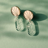 Elevate your style with these handcrafted Face Earrings: modern designs featuring round tops and dangling clear pieces etched with minimalist abstract faces, exuding artful sophistication on a green surface with soft shadows.