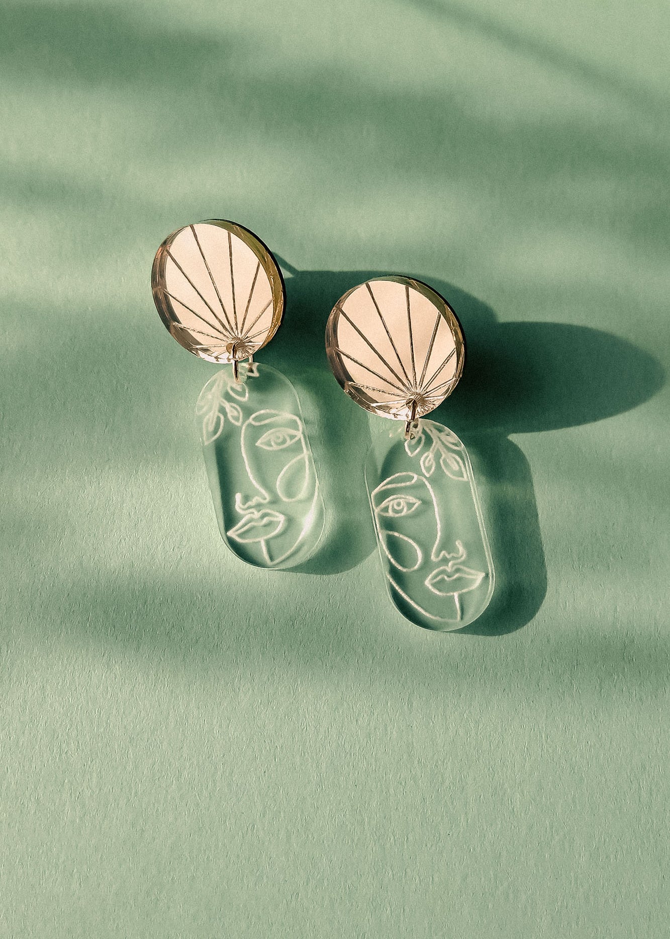 Elevate your style with these handcrafted Face Earrings: modern designs featuring round tops and dangling clear pieces etched with minimalist abstract faces, exuding artful sophistication on a green surface with soft shadows.