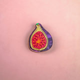 A handcrafted Fig Pin shaped like a halved fig, featuring a purple outer edge, gold outline, and vibrant red and yellow seeds, makes a lightweight accessory for any jacket or bag. Displayed on a pink background.