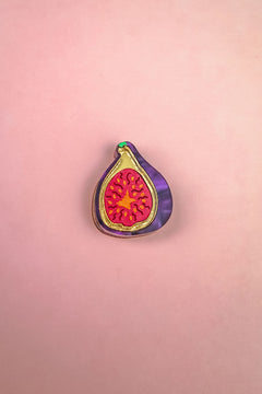 A handcrafted Fig Pin shaped like a halved fig, featuring a purple outer edge, gold outline, and vibrant red and yellow seeds, makes a lightweight accessory for any jacket or bag. Displayed on a pink background.