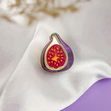 A handcrafted Fig Pin, shaped like a fig with a purple outline and red, seed-dotted center, rests on white satin fabric. A blurred beige plant sits in the corner, with a purple surface beneath this lightweight accessory.
