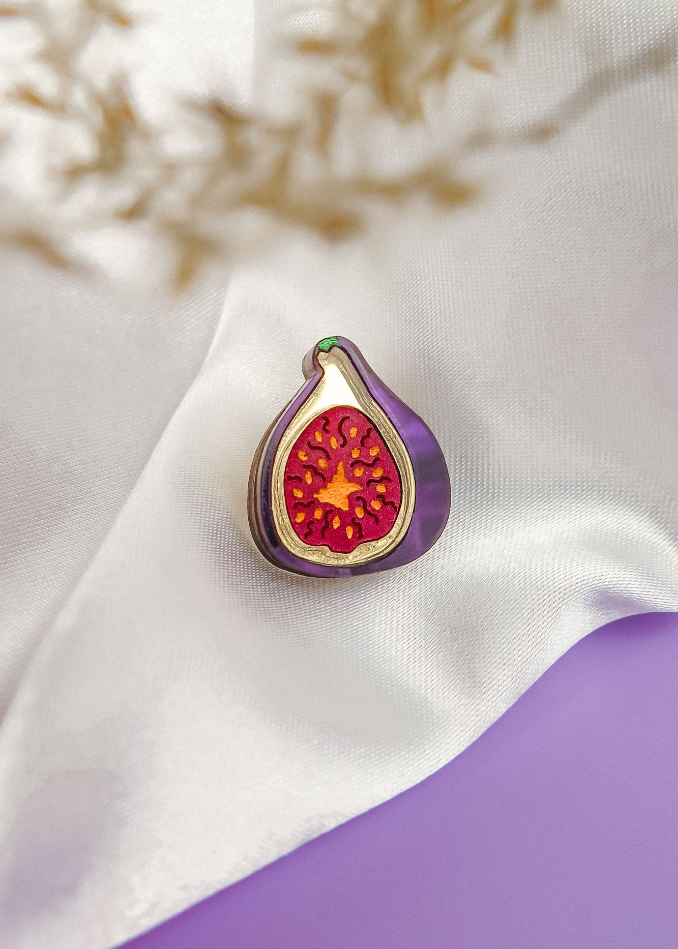 A handcrafted Fig Pin, shaped like a fig with a purple outline and red, seed-dotted center, rests on white satin fabric. A blurred beige plant sits in the corner, with a purple surface beneath this lightweight accessory.