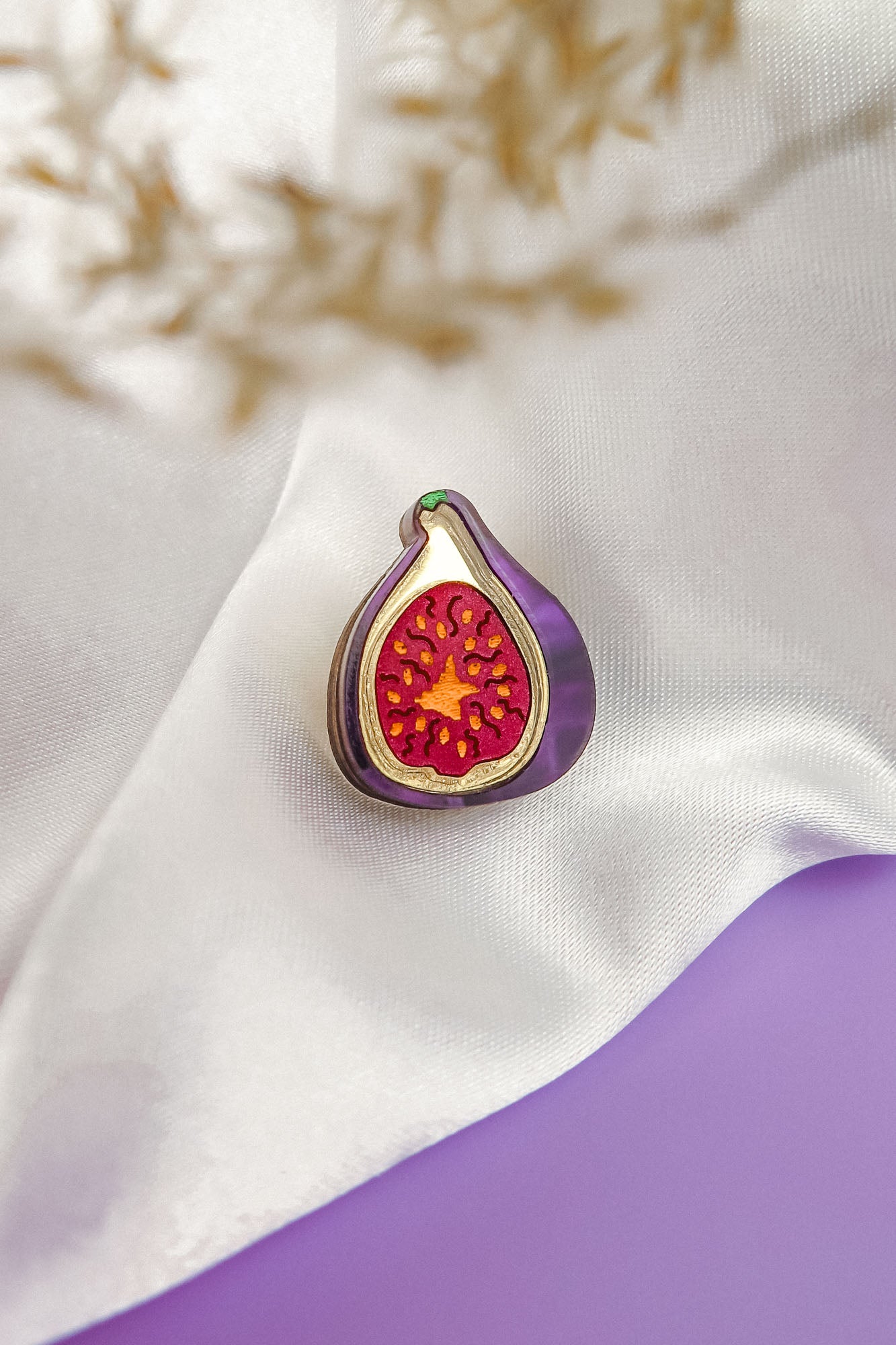 A handcrafted Fig Pin, shaped like a fig with a purple outline and red, seed-dotted center, rests on white satin fabric. A blurred beige plant sits in the corner, with a purple surface beneath this lightweight accessory.