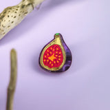 A colorful handcrafted Fig Pin with a red and yellow center and purple outer edge rests on a light purple surface, framed by two pieces of textured driftwood. This lightweight accessory adds charm to any collection.