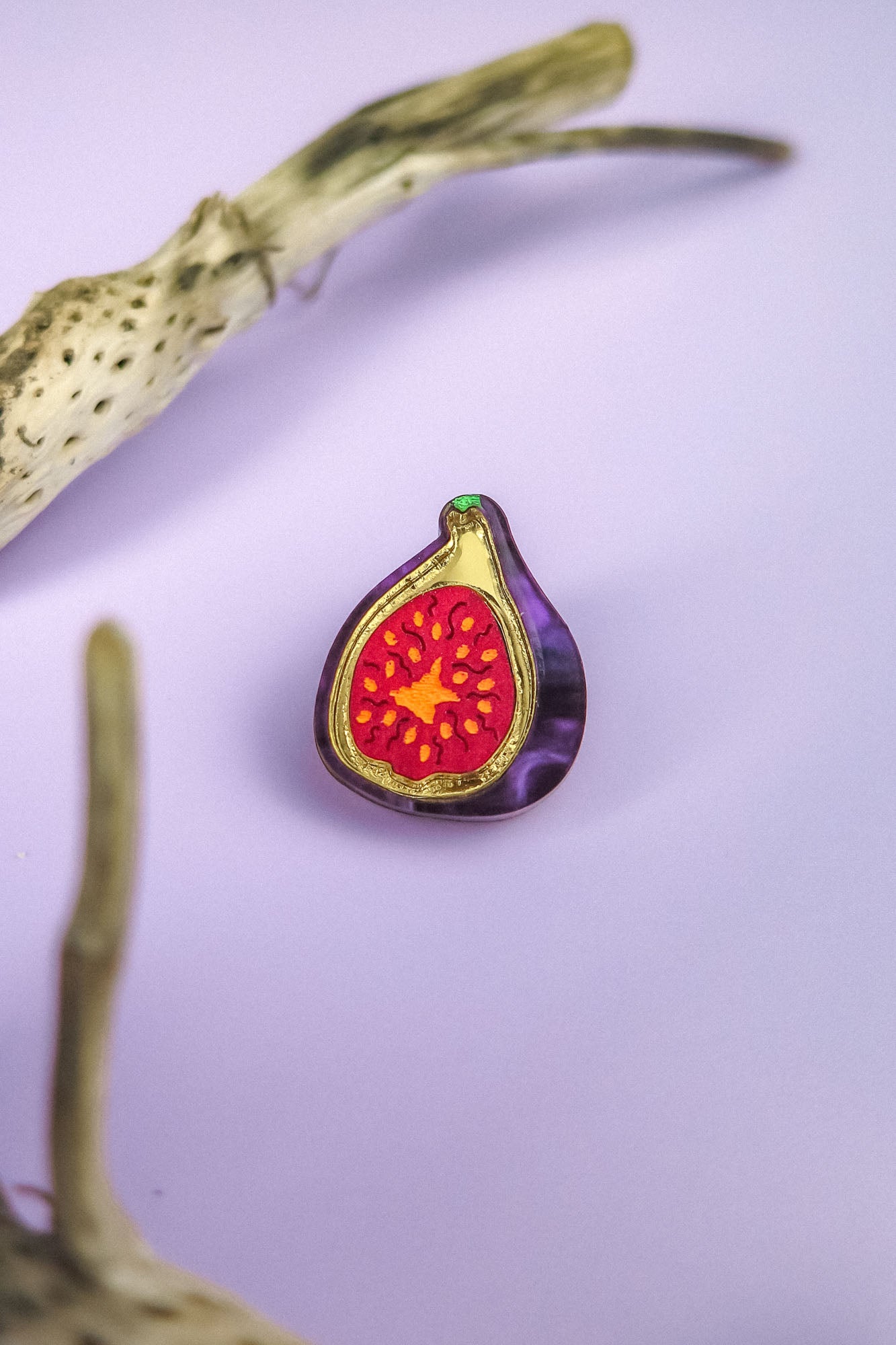 A colorful handcrafted Fig Pin with a red and yellow center and purple outer edge rests on a light purple surface, framed by two pieces of textured driftwood. This lightweight accessory adds charm to any collection.