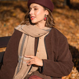 A woman wearing a brown beret, a brown coat, tan scarf, and handcrafted earrings sits outside on a bench surrounded by autumn leaves, looking to the side and smiling softly in the sunlight.