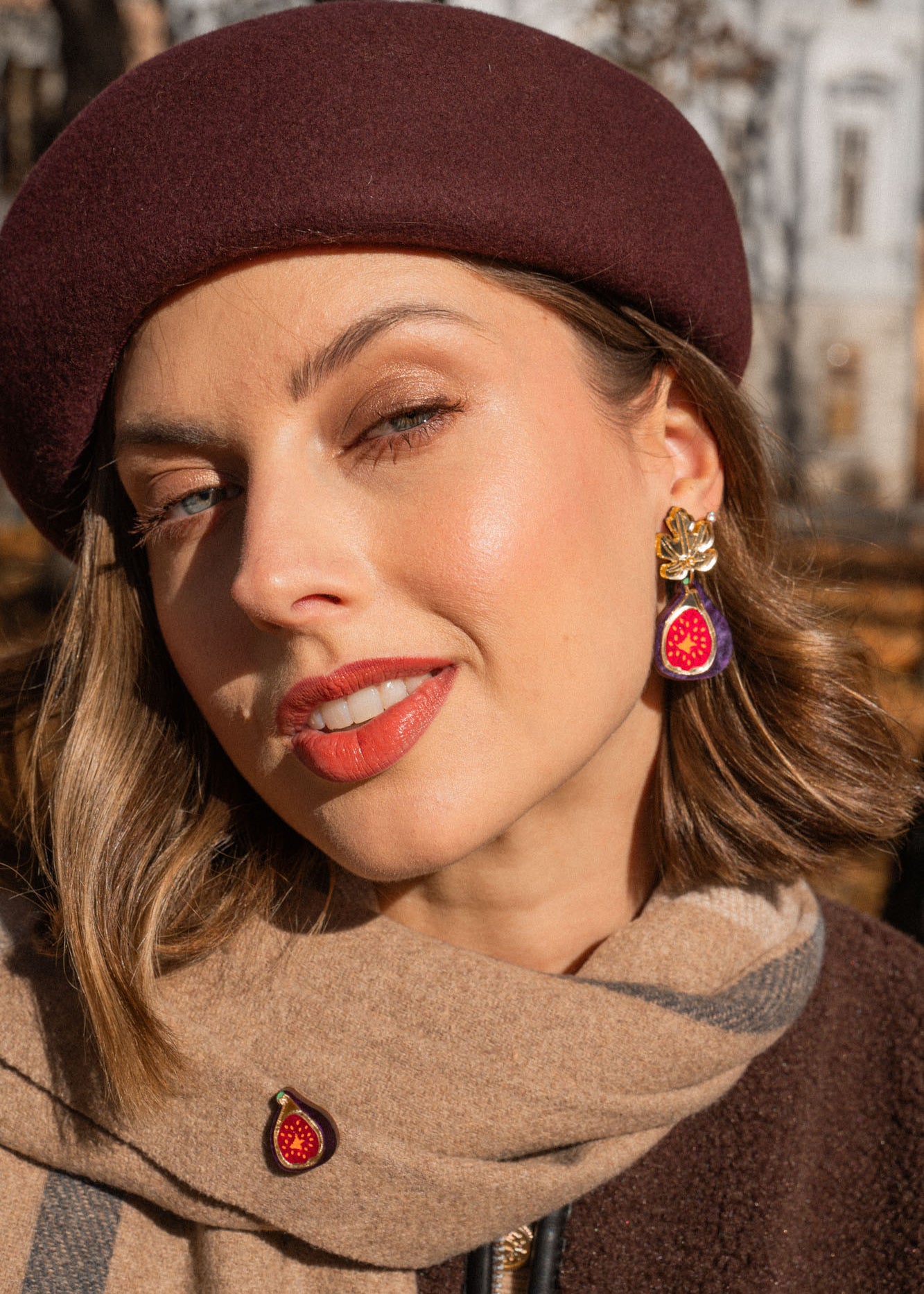 A woman wearing a burgundy beret and a beige scarf poses outdoors. She wears red lipstick, handcrafted fig earrings, and a matching pin. Autumn trees and a building create a scenic background.