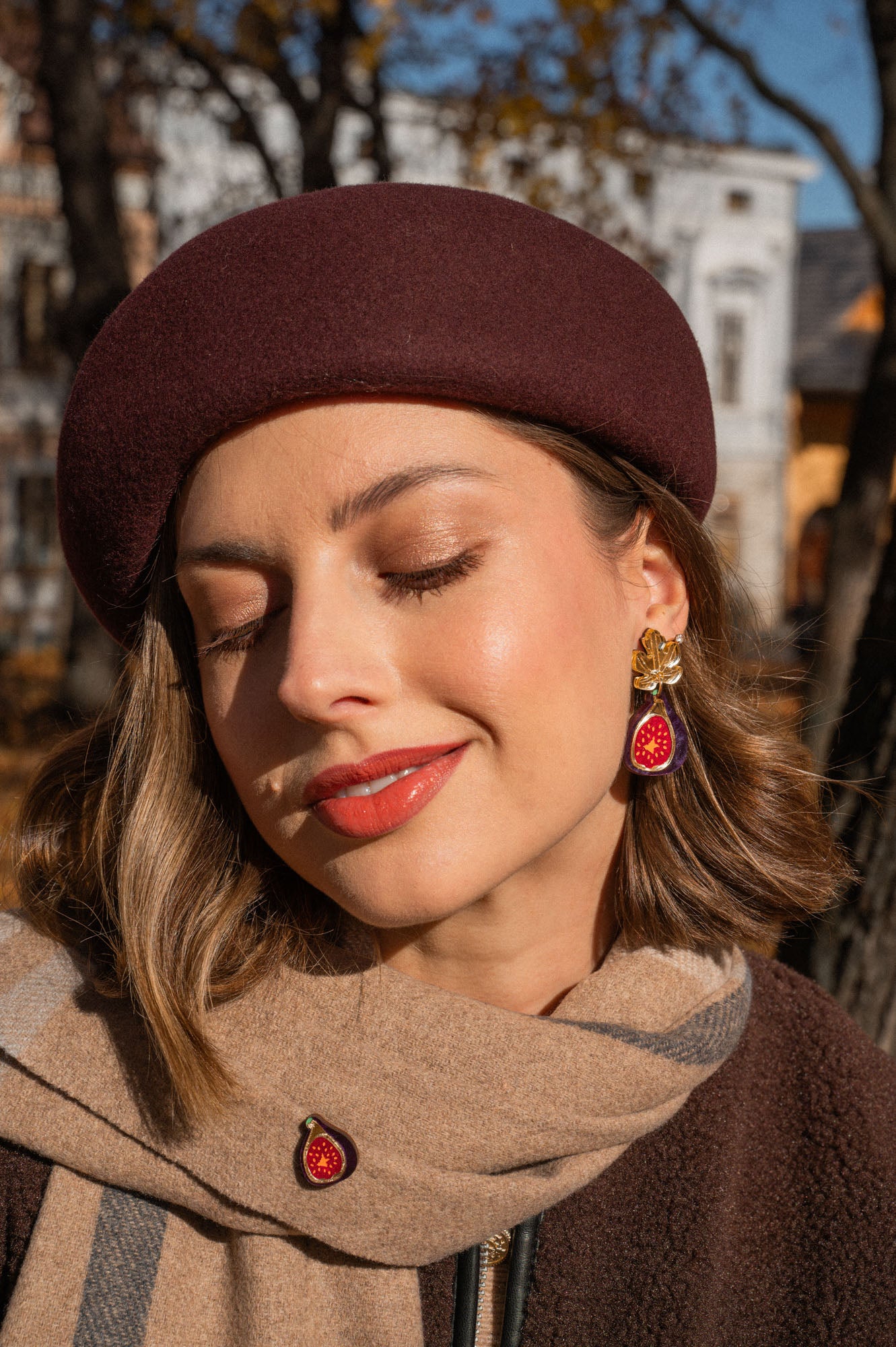A woman outdoors smiles gently with her eyes closed. She wears a burgundy beret, a tan scarf with a fig brooch, and handcrafted fig earrings accented with gold mirror leaves. Autumn trees and buildings are blurred in the background.