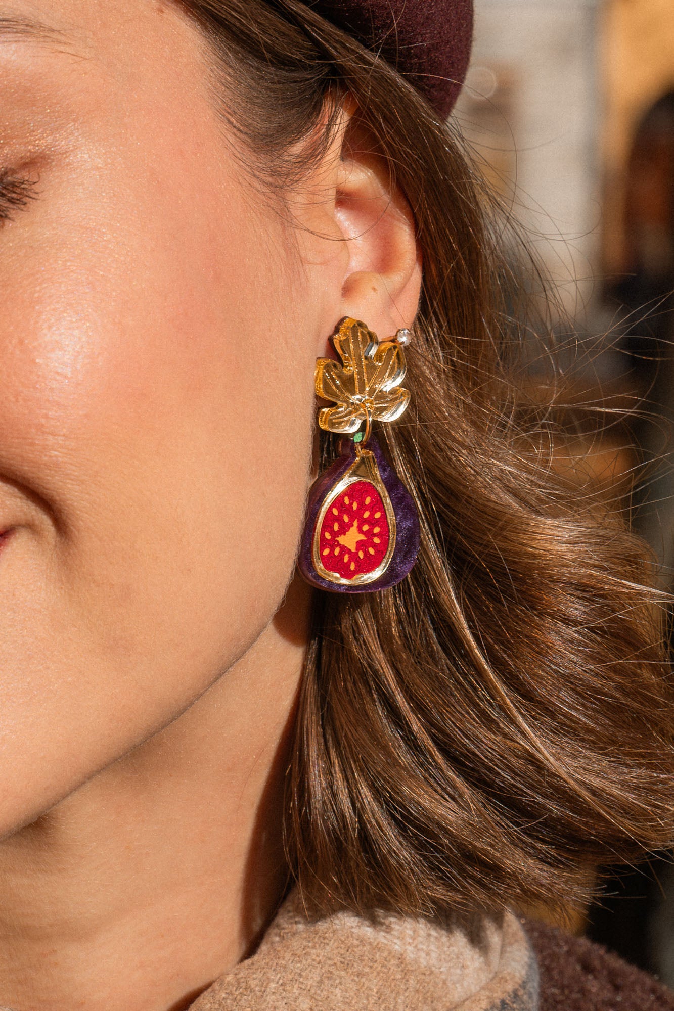 A woman outdoors smiles gently with her eyes closed. She wears a burgundy beret, a tan scarf with a fig brooch, and handcrafted fig earrings accented with gold mirror leaves. Autumn trees and buildings are blurred in the background.