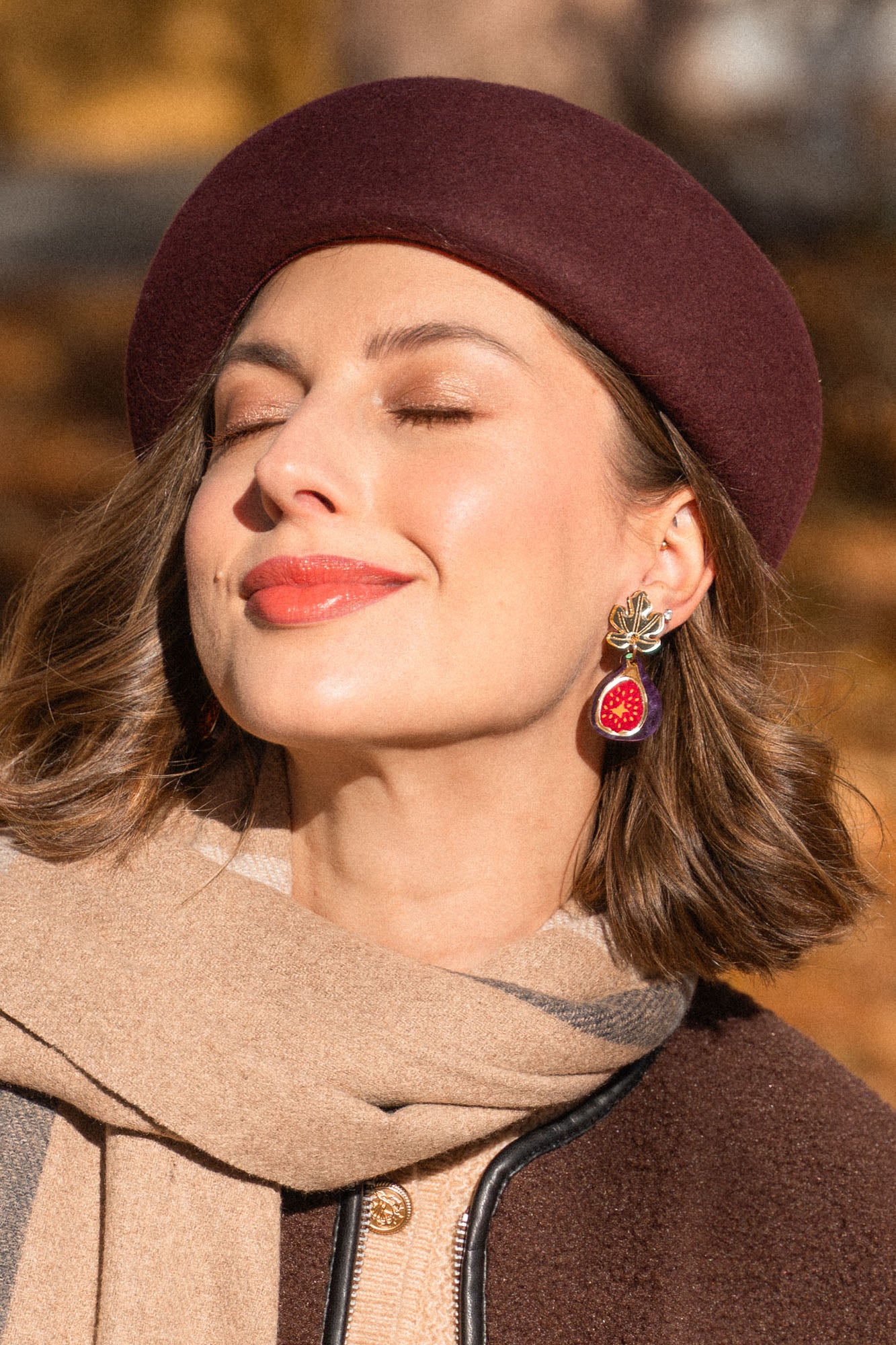 A woman with light brown hair wearing a burgundy beret, beige scarf, and handcrafted fig earrings smiles with her eyes closed in warm sunlight, with a blurred autumn background.