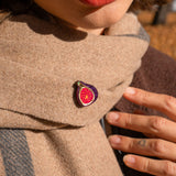 A person wearing red lipstick and a beige scarf with a handcrafted Fig Pin, a unique accessory shaped like a fig; their hand rests on the scarf, showing neat nails and a small finger tattoo.