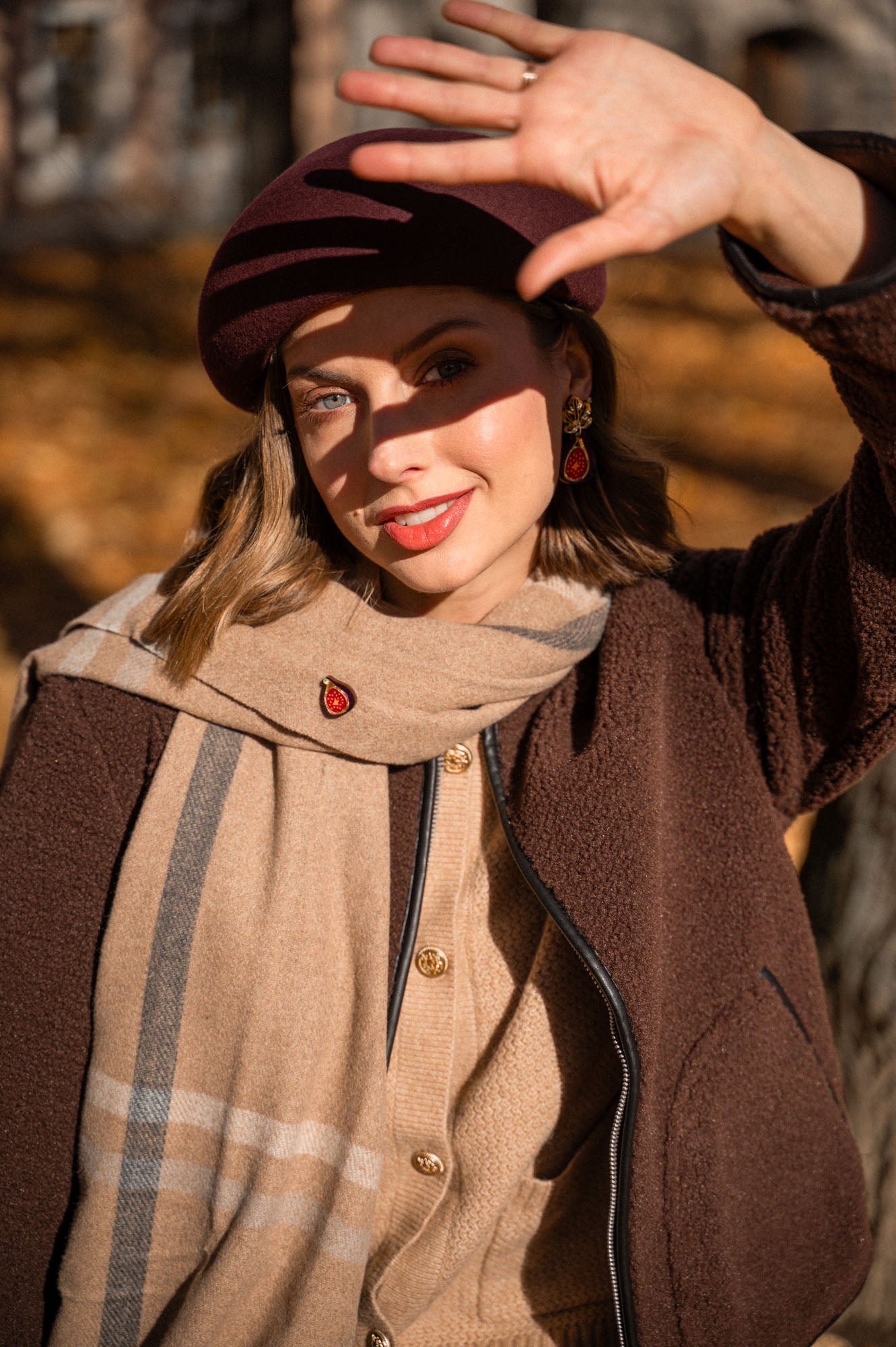 A woman in a brown beret and coat smiles at the camera, holding one hand up to shield her eyes from the sunlight. She wears a beige scarf, red earrings, and a unique accessory—a handcrafted Fig Pin—with autumn leaves blurred in the background.