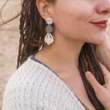 A woman with closed eyes and long brown dreadlocks wears a beige sweater and large handcrafted boho earrings, standing outdoors on a sandy beach. She smiles softly and touches her chest with one hand.